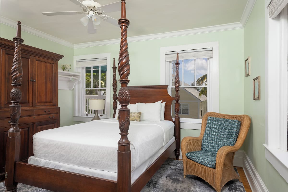 A four-poster bed with a wooden headboard, a wicker chair, and a ceiling fan in a room with light green walls and white trim.