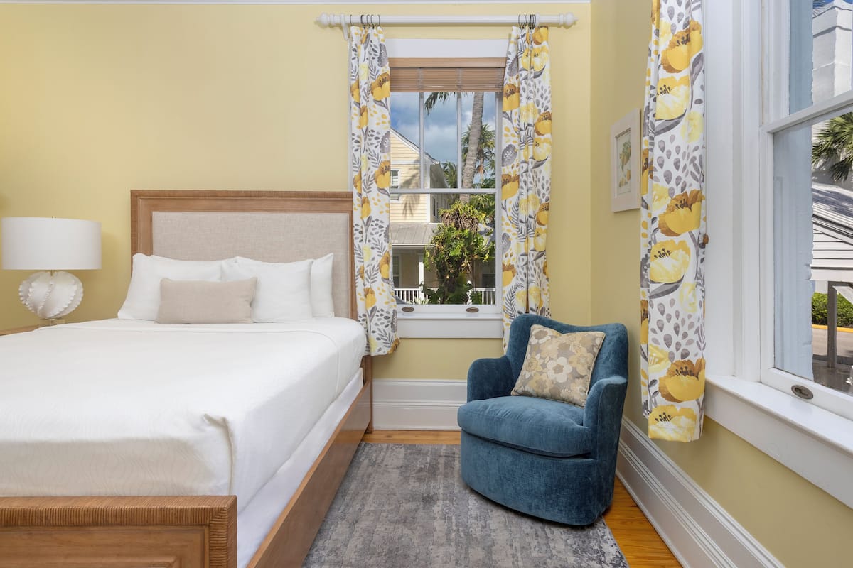 A bedroom with a bed, a chair, a window with curtains, and a lamp.