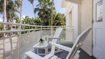 A balcony with white chairs and a small table, overlooking a garden with palm trees.