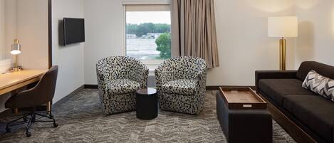 A hotel room with a desk, two patterned armchairs, a sofa, a small ottoman, a TV, and a window with a view of a bridge and trees.