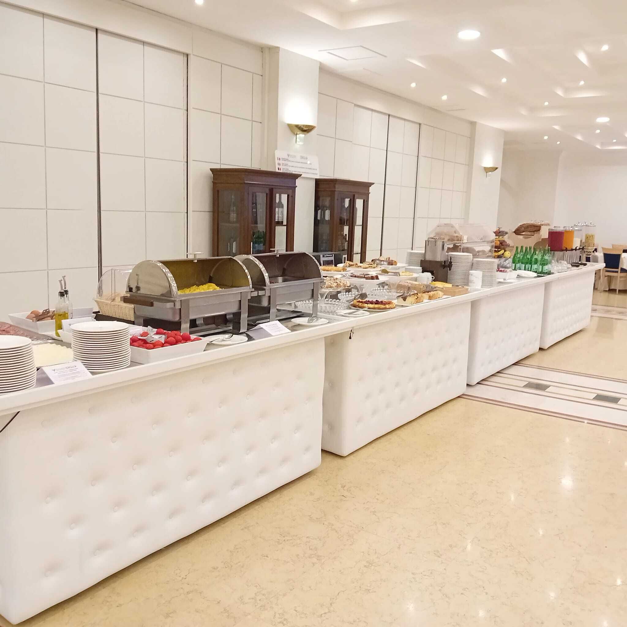 Disposição de buffet com diversos alimentos e bebidas, uma parede revestida de azulejos e um armário de madeira ao fundo.