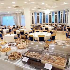 A banquet hall with round tables and blue chairs, featuring a buffet with various baked goods and pastries.