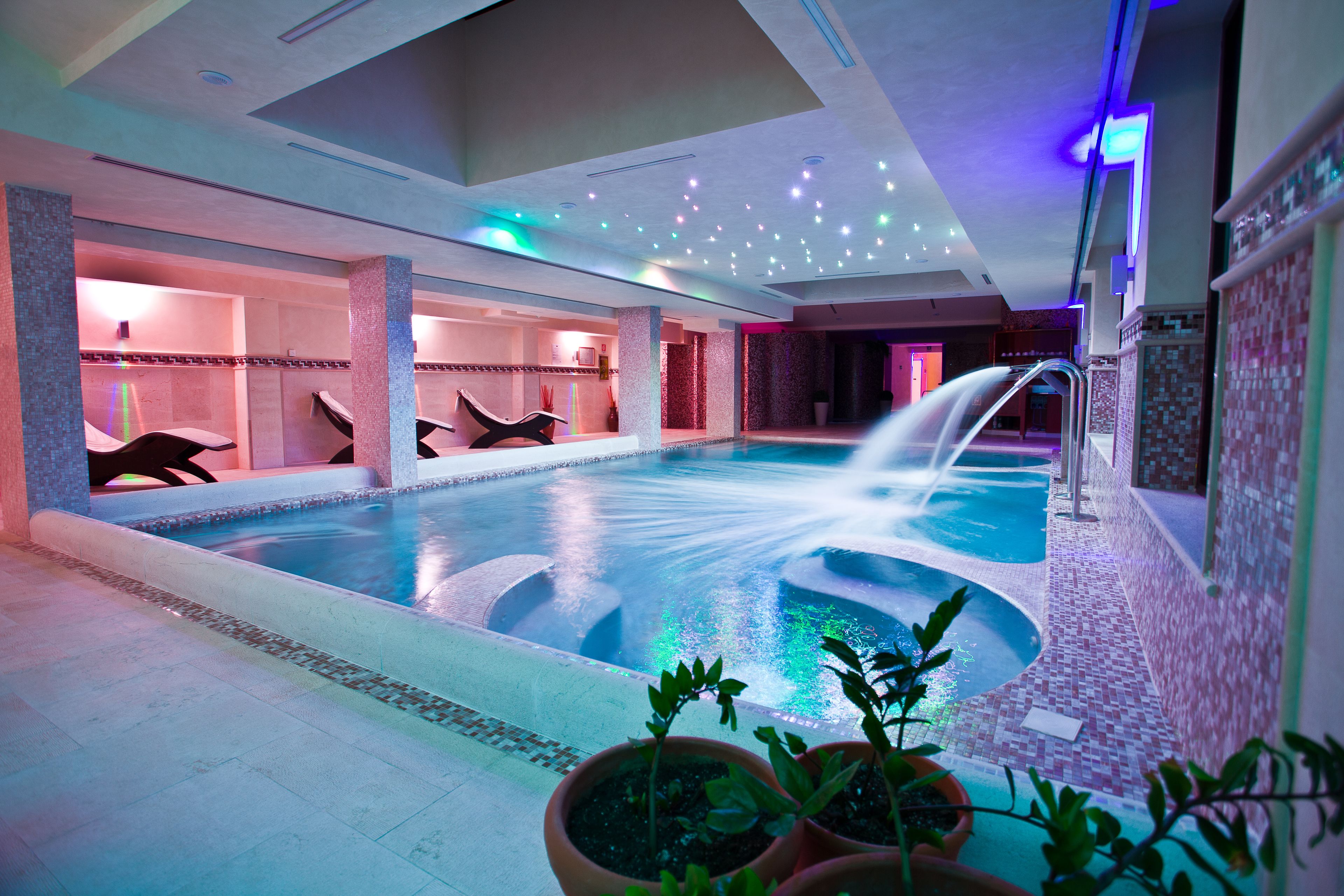 An indoor swimming pool with a water fountain, tiled walls, and potted plants.