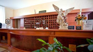 A reception desk with a 'RECEPTION' sign, a statue of an angel, a wine rack, and a vase with flowers.