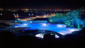 A swimming pool with a curved edge, surrounded by lounge chairs and a view of a cityscape at night. - Palace Hotel San Michele (Monte Sant'Angelo)