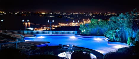 Una piscina con borde curvo, rodeada de sillones de descanso y vista a un paisaje urbano nocturno.