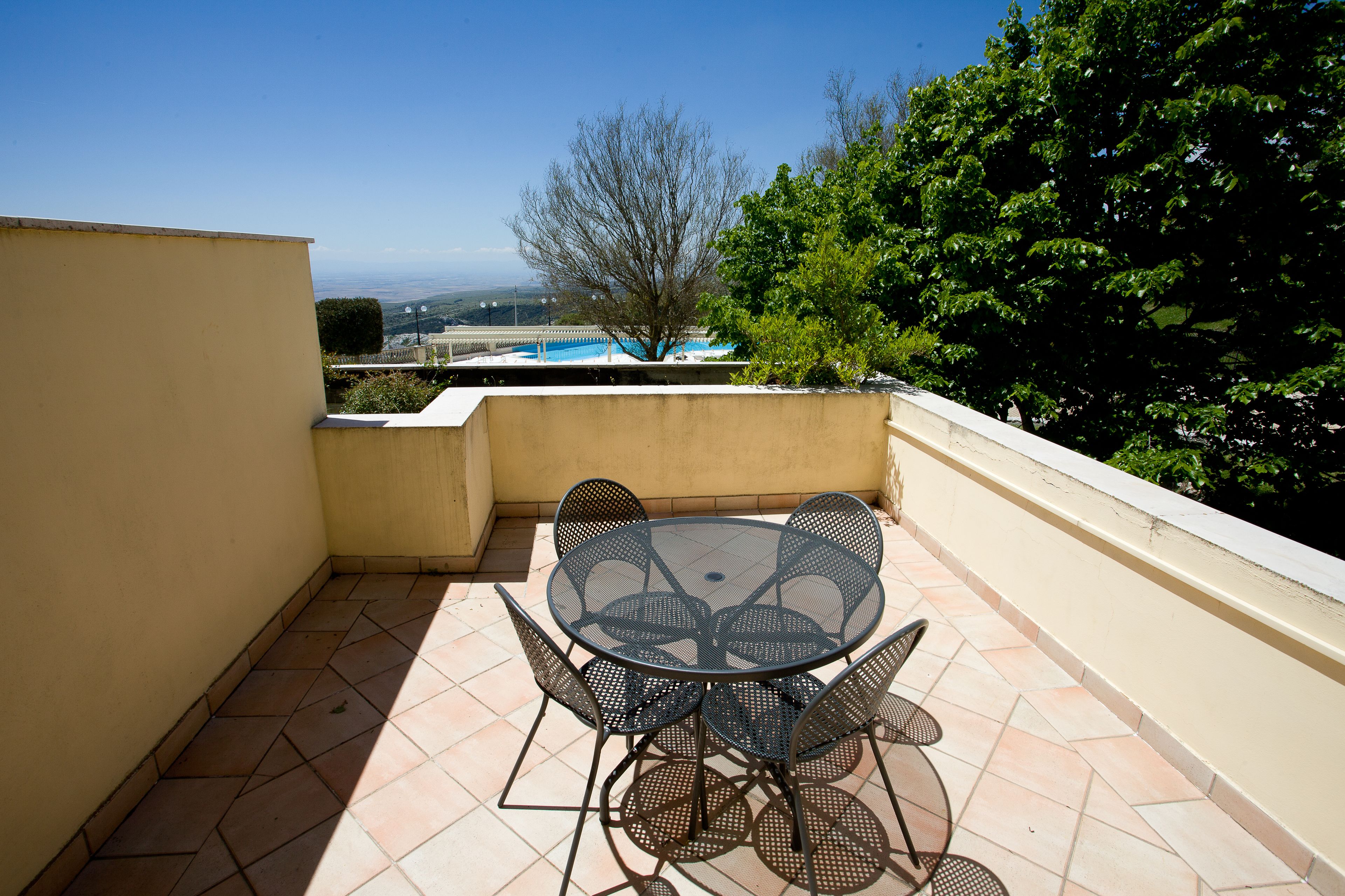 A small outdoor patio with a round table and chairs, a tiled floor, and a view of a swimming pool and trees in the background.