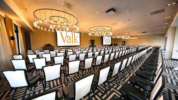 A conference room with rows of chairs, a large screen displaying 'Valk', and modern chandeliers.