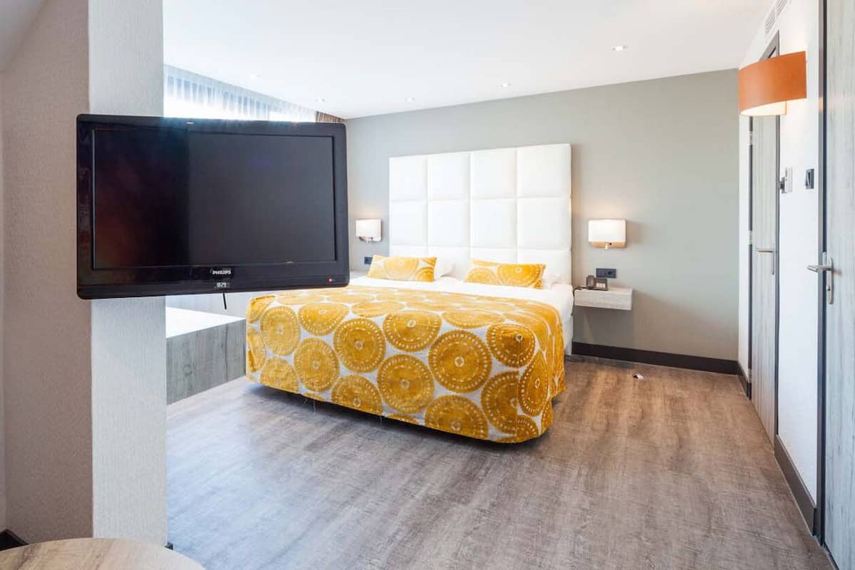 Ein Hotelzimmer mit einem großen Bett, einem an der Wand montierten Fernseher und einem Nachttisch mit einer Lampe.