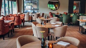 Een restaurant met ronde tafels en stoelen, grote ramen en een spiegel aan de muur.