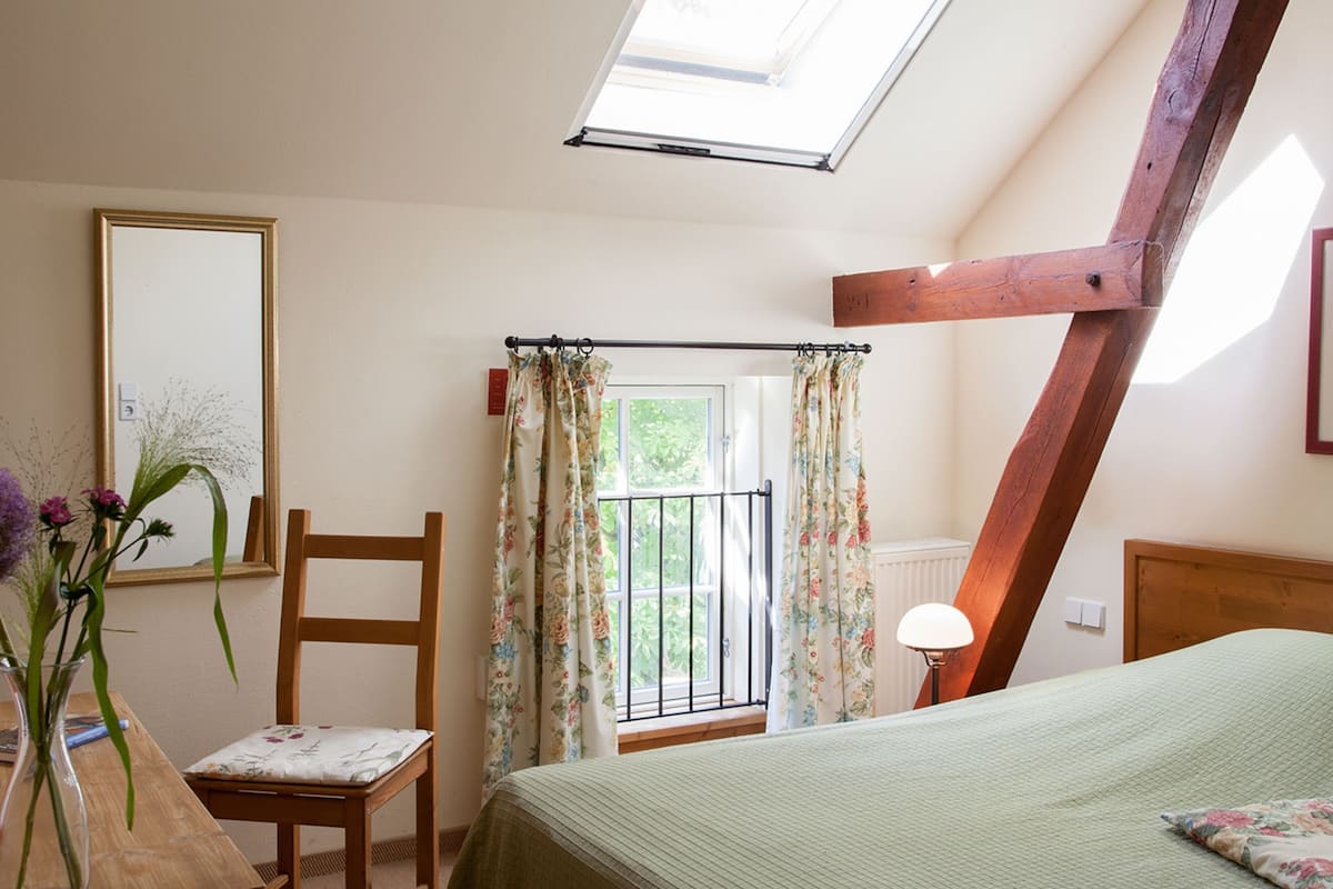 Une chambre avec un puits de lumière, un lit, une chaise, un bureau et une fenêtre avec des rideaux.