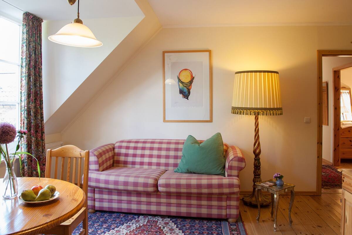 Un salon chaleureux avec un canapé à carreaux, une table garnie de fruits, une lampe et une œuvre d’art encadrée accrochée au mur.