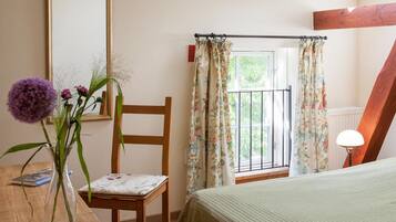 Un dormitorio con una cama, una silla, una mesa con un jarrón de flores, una ventana con cortinas y una lámpara.
