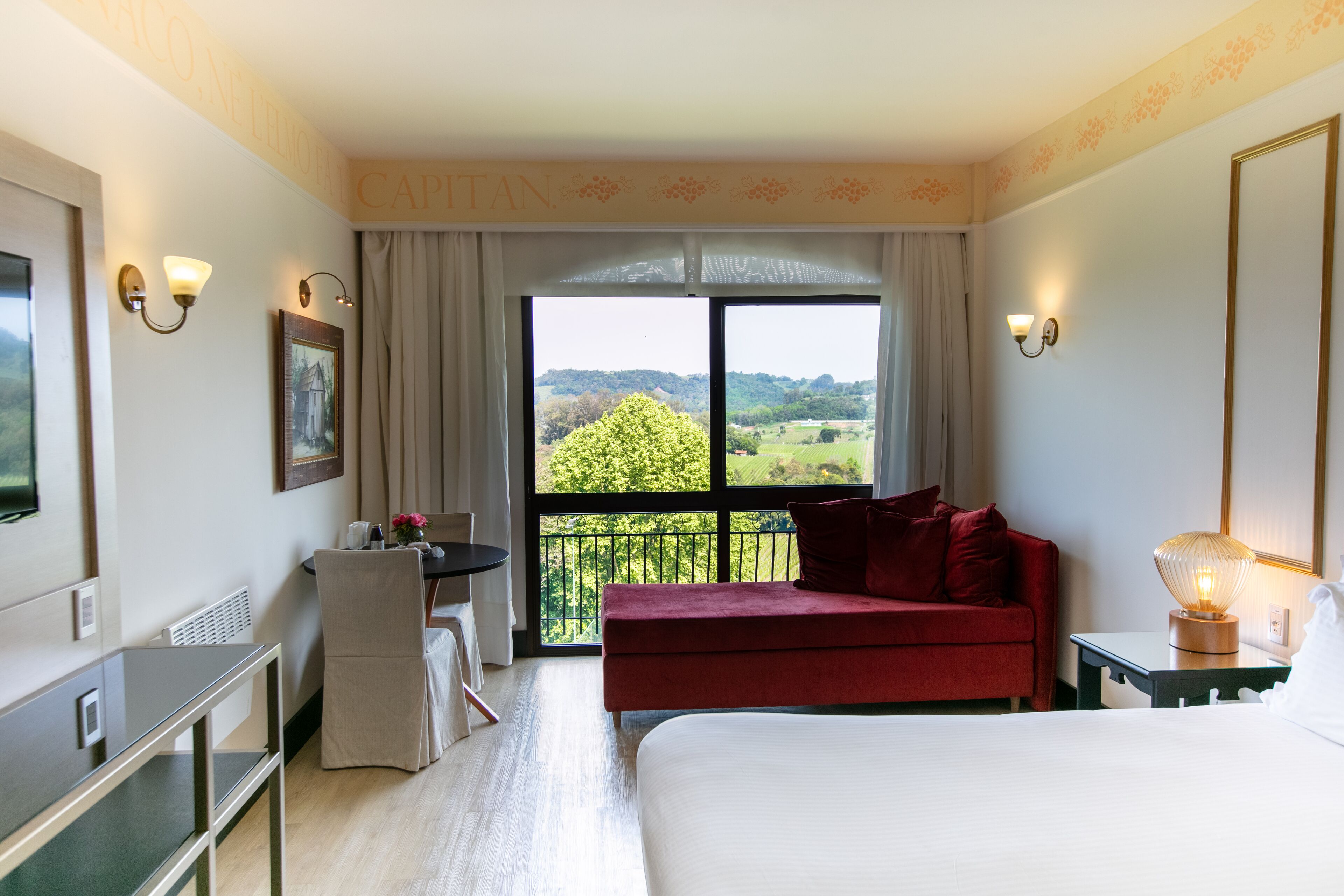 A hotel room with a large bed, a red sofa, a desk with a chair, a small table with a lamp, and a large window offering a view of greenery.