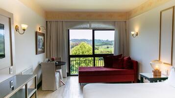 A hotel room with a large bed, a red sofa, a desk with a chair, a small table with a lamp, and a large window offering a view of greenery.