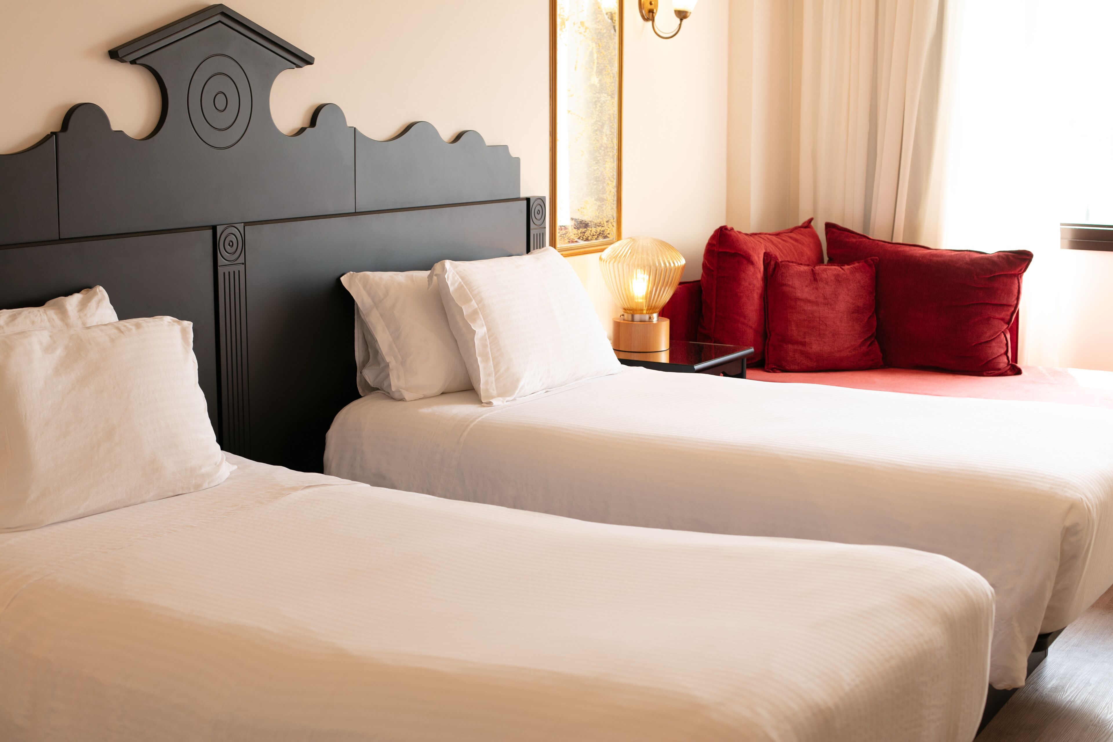 Dos camas con sábanas blancas, cabecero oscuro, almohada roja y una lámpara fijada a la pared.