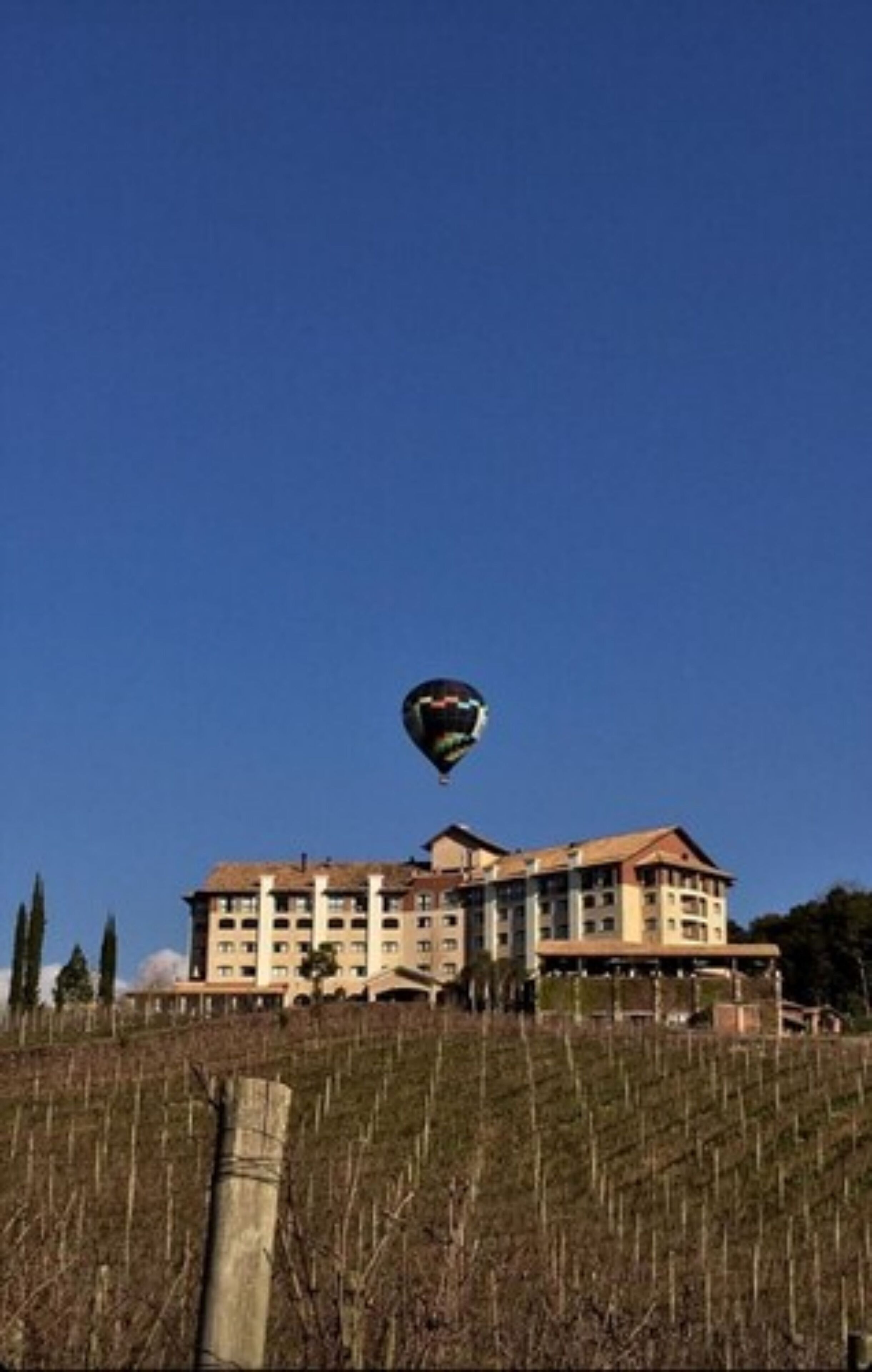 A hot air balloon flying over a large building with multiple floors and a sloped roof.