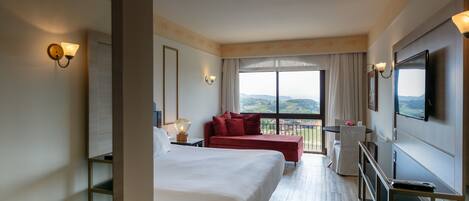 Habitación de hotel con una cama grande, un sofá rojo, un televisor y un balcón con vistas.