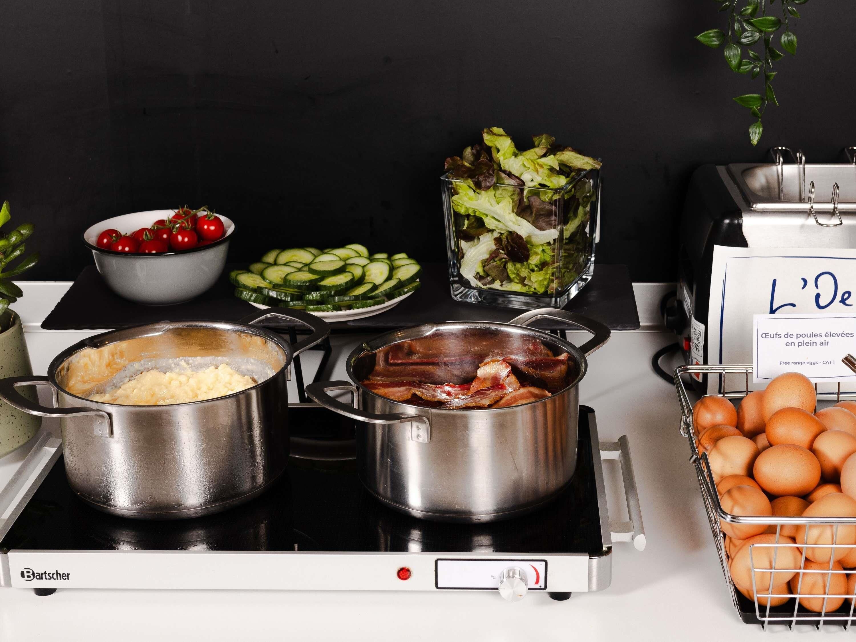 A kitchen with two pots on a stove, a bowl of cherries, a plate of sliced cucumbers, a salad, and a basket of eggs.