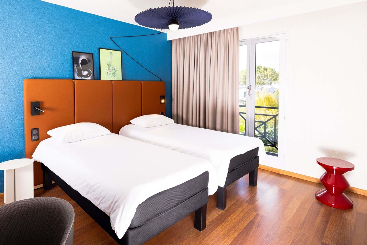A hotel room with a double bed, a small desk, a chair, a red side table, and a balcony with a view.