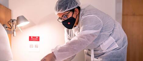 A person in a white uniform and hairnet cleaning a bed in a hotel room.