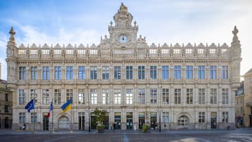 Um edifício grande e ornamentado com uma torre do relógio, várias bandeiras e um pátio pavimentado.