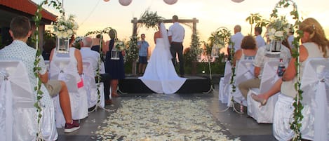Una ceremonia de boda con invitados sentados en sillas dispuestas en filas, mirando hacia un escenario donde una pareja se encuentra bajo un arco floral.