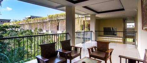 A spacious balcony with wooden furniture and a glass table, overlooking a lush garden and a building with vines.