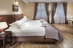 Ein Hotelzimmer mit einem großen Bett, einem Nachttisch mit Lampe, einem Sessel und einem Fenster mit durchsichtigen Vorhängen.