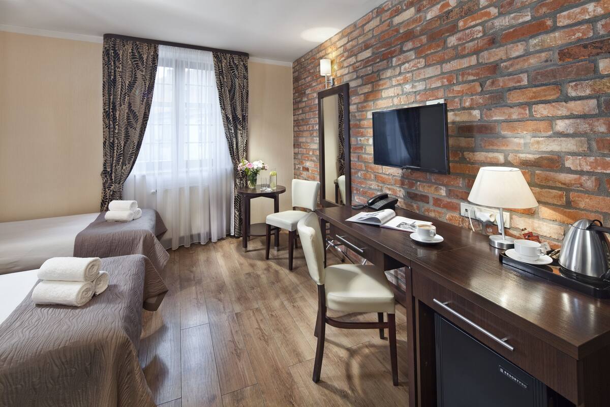 Habitación de hotel con dos camas, un escritorio con lámpara, cafetera y televisor fijado a una pared de ladrillo.