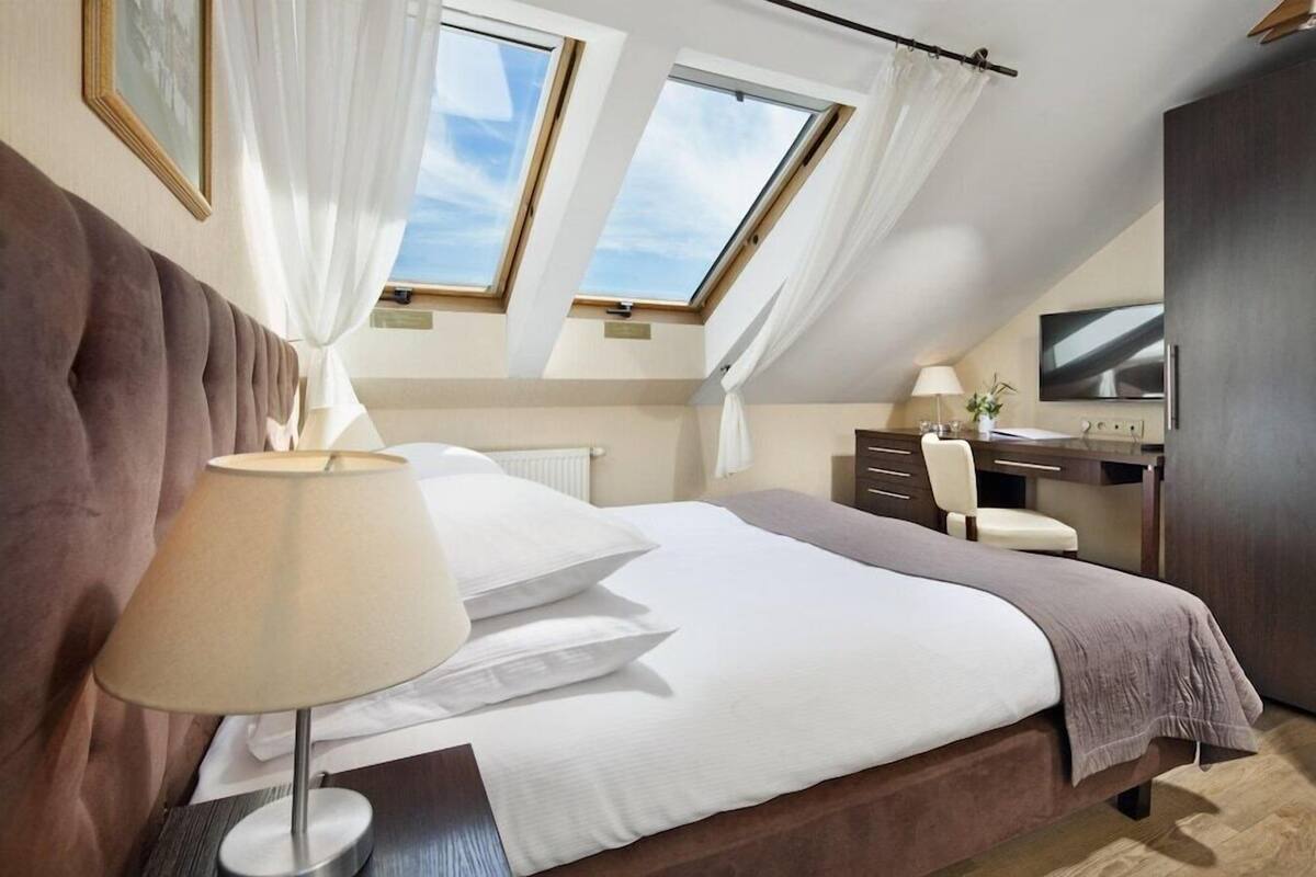 Un dormitorio con una cama grande, un escritorio, una silla y un televisor. Hay tragaluces y cortinas.
