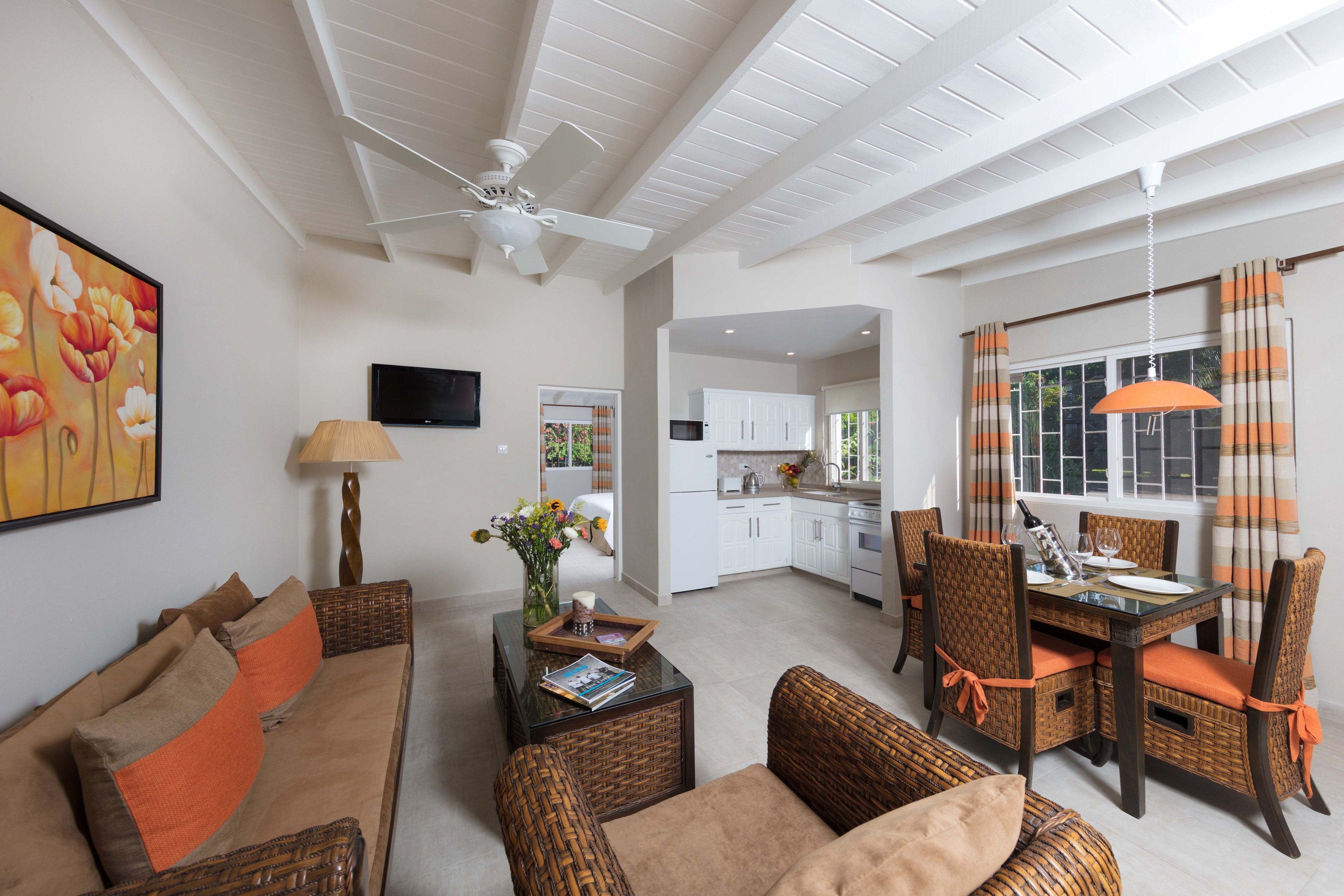 A spacious living area with a sofa, coffee table, and dining table set. A ceiling fan, kitchen, and window with curtains are visible.