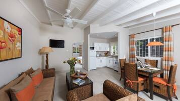 A spacious living area with a sofa, coffee table, and dining table set. A ceiling fan, kitchen, and window with curtains are visible.