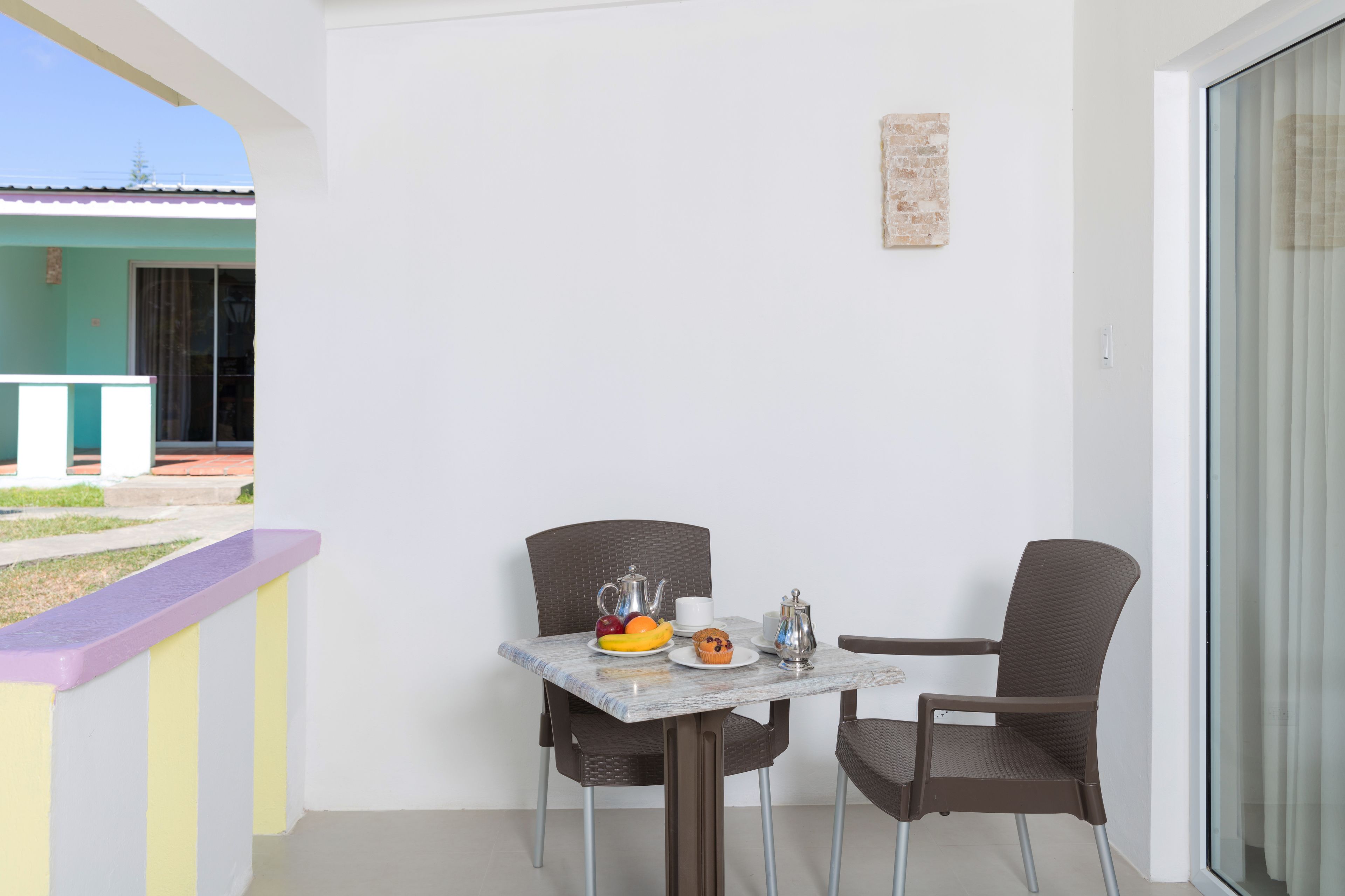 A balcony with a table and two chairs, a small table with fruits and a teapot, and a view of a building.