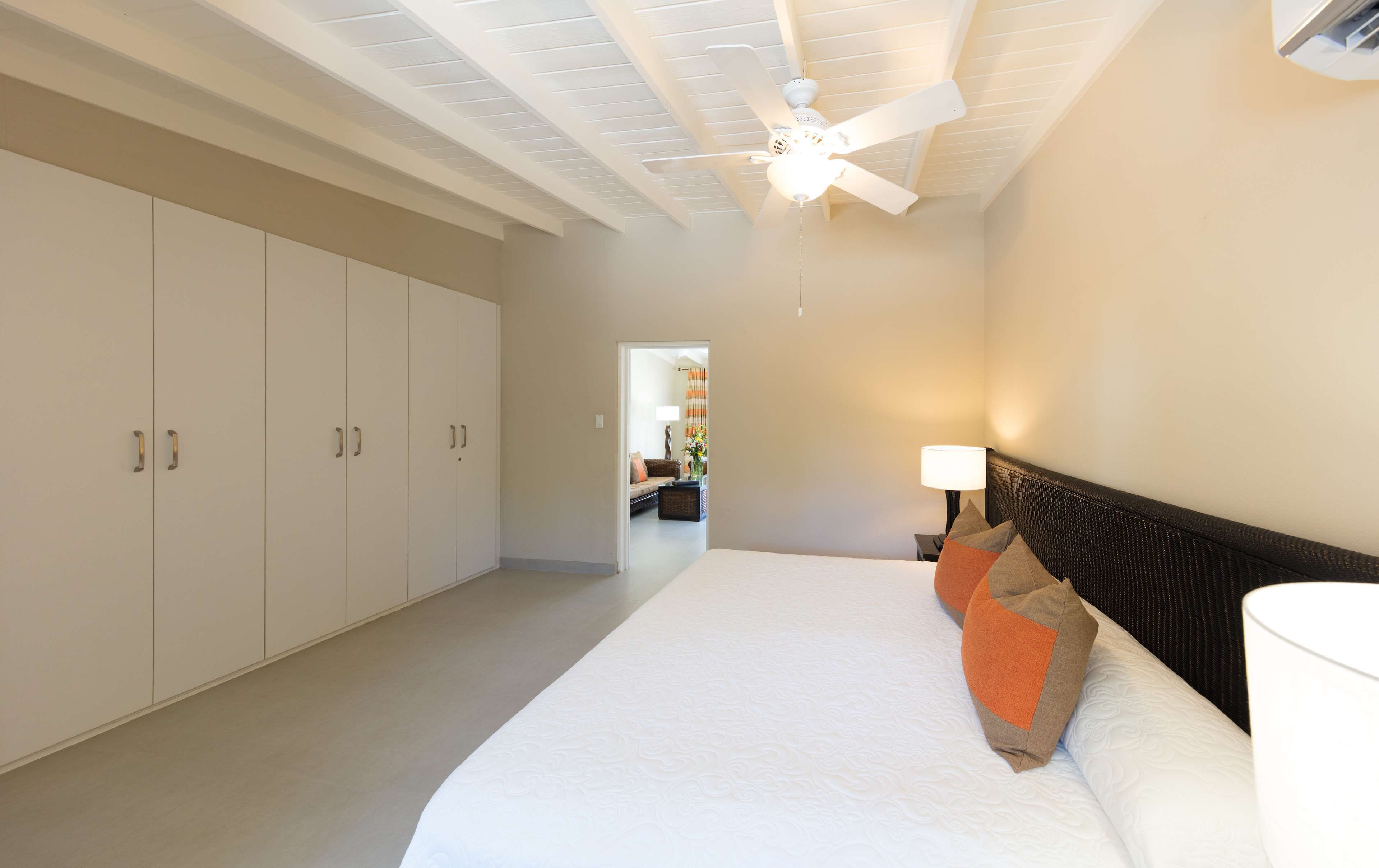 Un dormitorio con cama, ventilador de techo, mesita de noche con lámpara y vista a un salón a través de una puerta abierta.