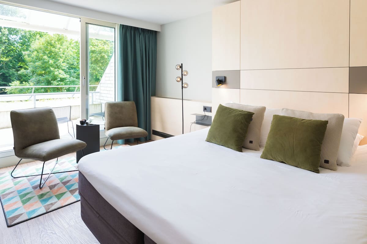 A hotel room with a large bed, two armchairs, a small table, and a view of greenery outside.