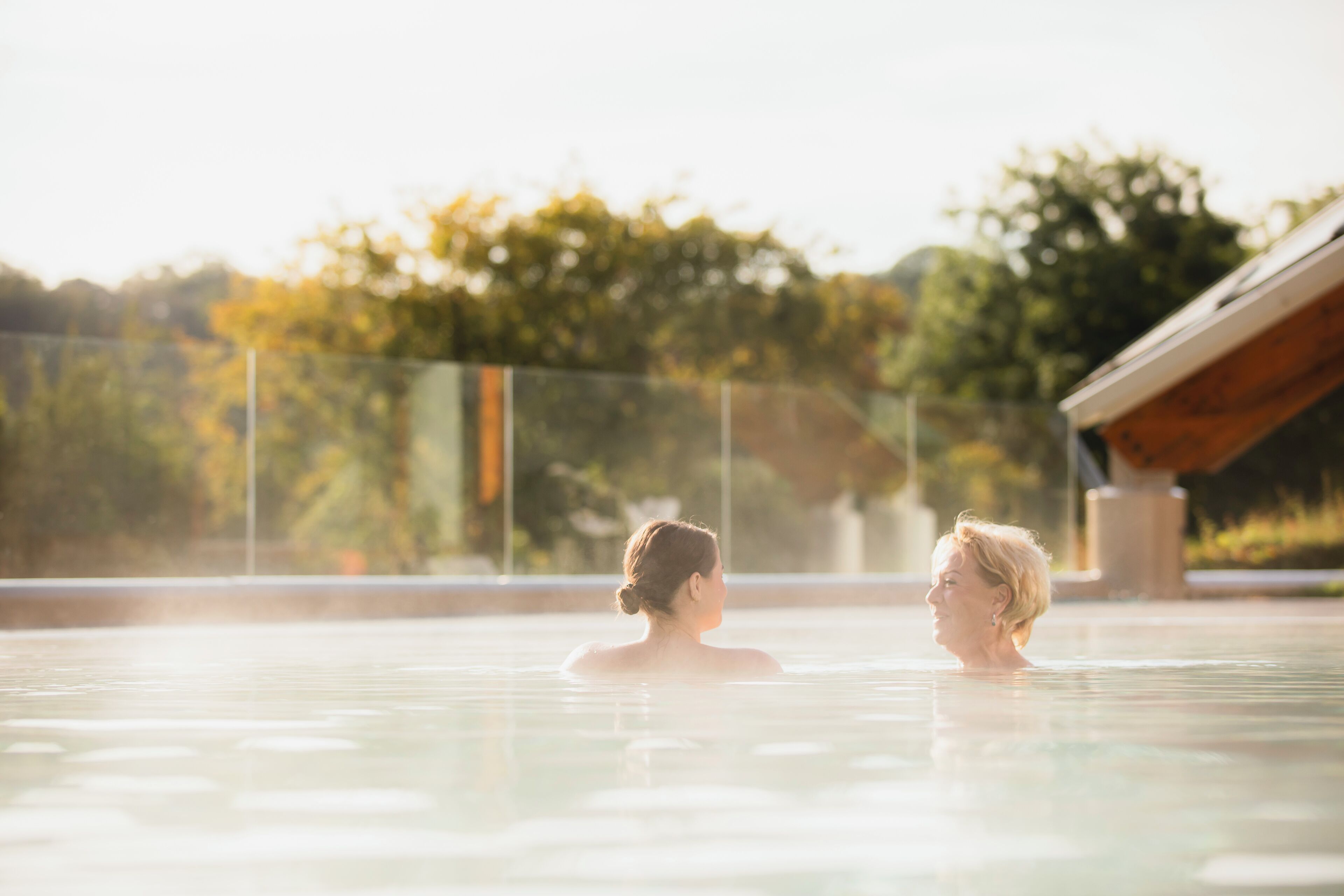 Zwei Personen in einem Pool mit Blick auf Bäume und ein Gebäude im Hintergrund.