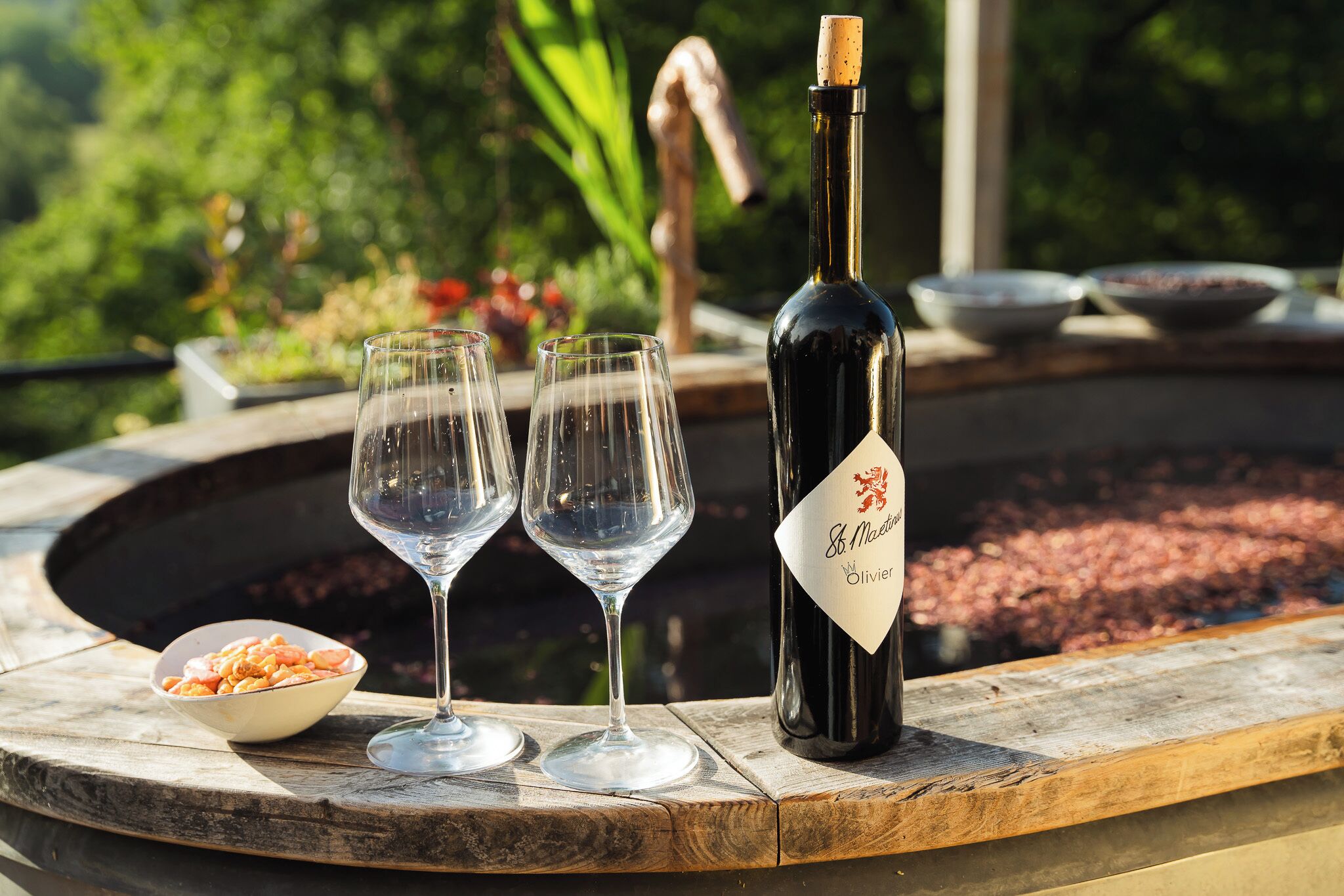 A bottle of wine, two glasses, and a bowl of shrimp on a wooden table outdoors.
