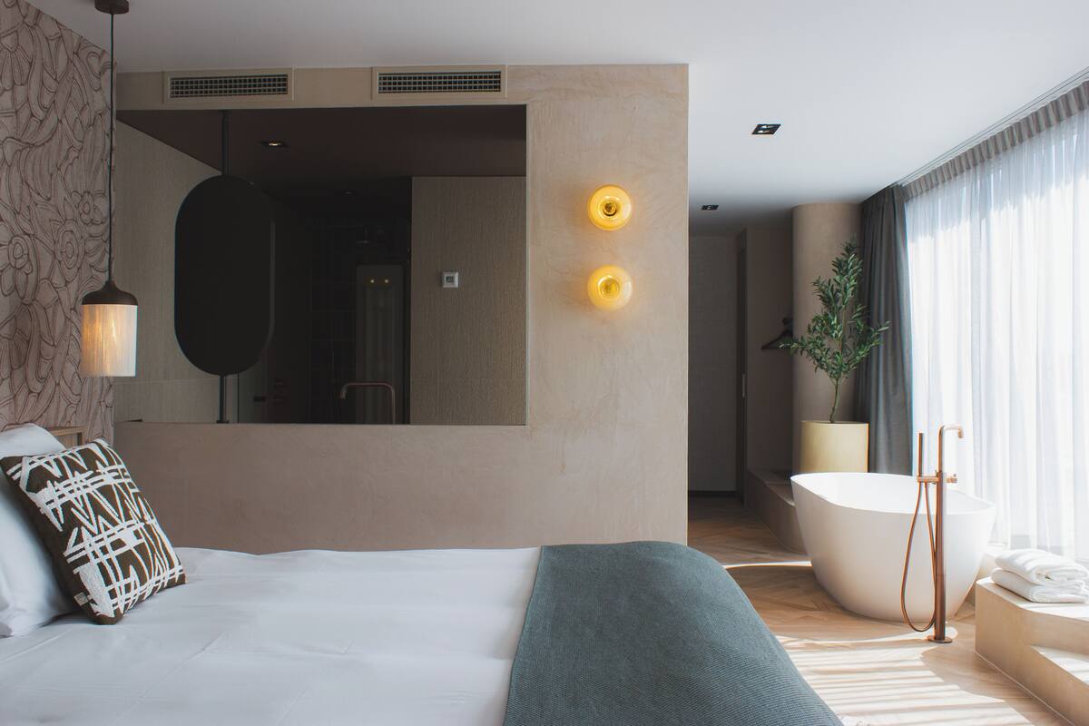 A modern hotel room with a large bed, a sitting area, a bathtub, and a large mirror.