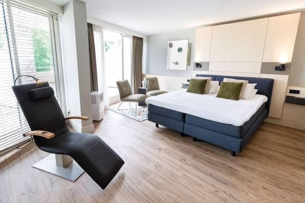 A modern bedroom with a large bed, a reclining chair, a desk, and a view of the outdoors.