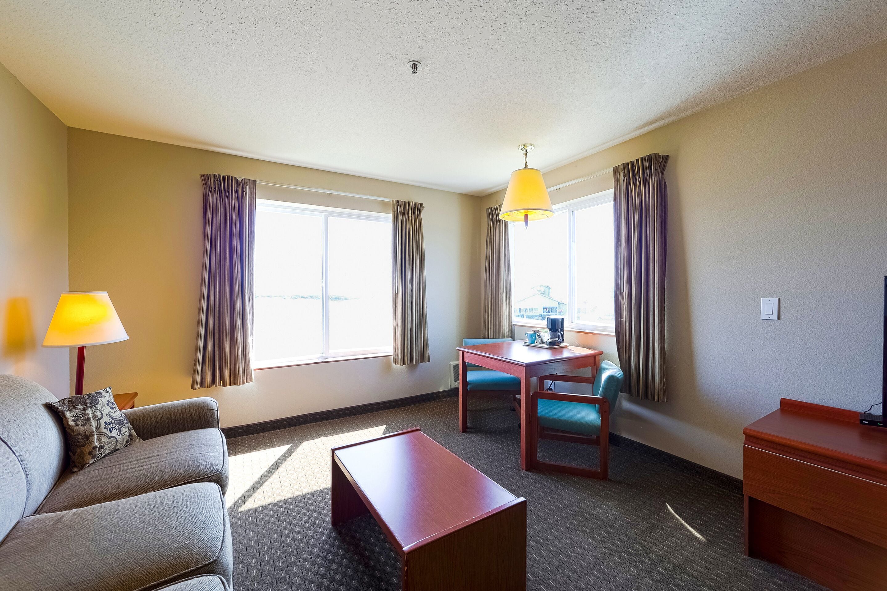 Habitación de hotel con sofá, una mesita, un escritorio y una silla. Hay ventanas con cortinas, una lámpara y una luz en el techo.