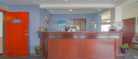 Una recepción con detalles de madera, una pared azul con el nombre "Silver Bay Lodge" y un logotipo, una puerta roja cerrada y dos plantas en macetas sobre pequeñas mesas.