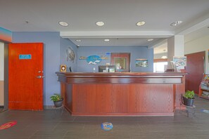 Balcão de recepção com acabamento em madeira, parede azul com o nome "Silver Bay Lodge" e um logotipo, porta vermelha fechada e duas plantas em vasos sobre pequenas mesas.