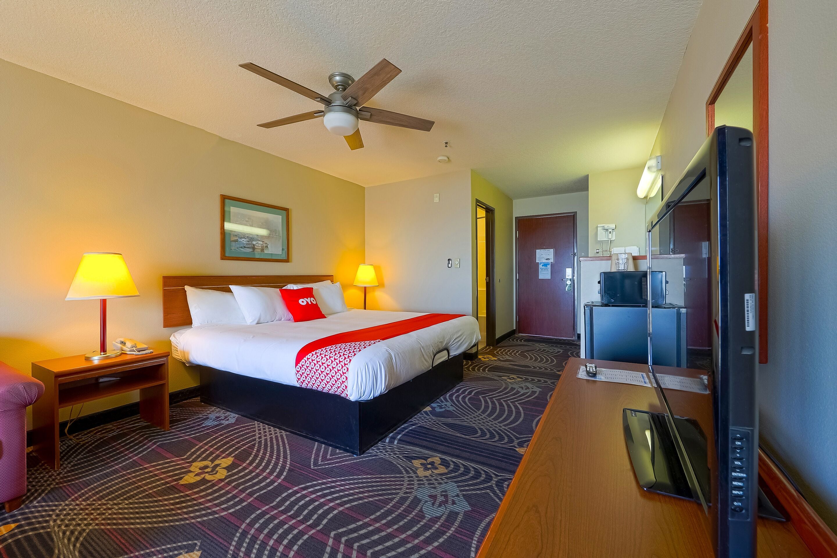 Quarto de hotel com cama, duas luminárias de cabeceira, ventoinha no teto, escrivaninha com televisão e tapete estampado.