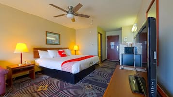 A hotel room with a bed, two bedside lamps, a ceiling fan, a desk with a TV, and a patterned carpet.