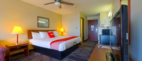 A hotel room with a bed, two bedside lamps, a ceiling fan, a desk with a TV, and a patterned carpet.