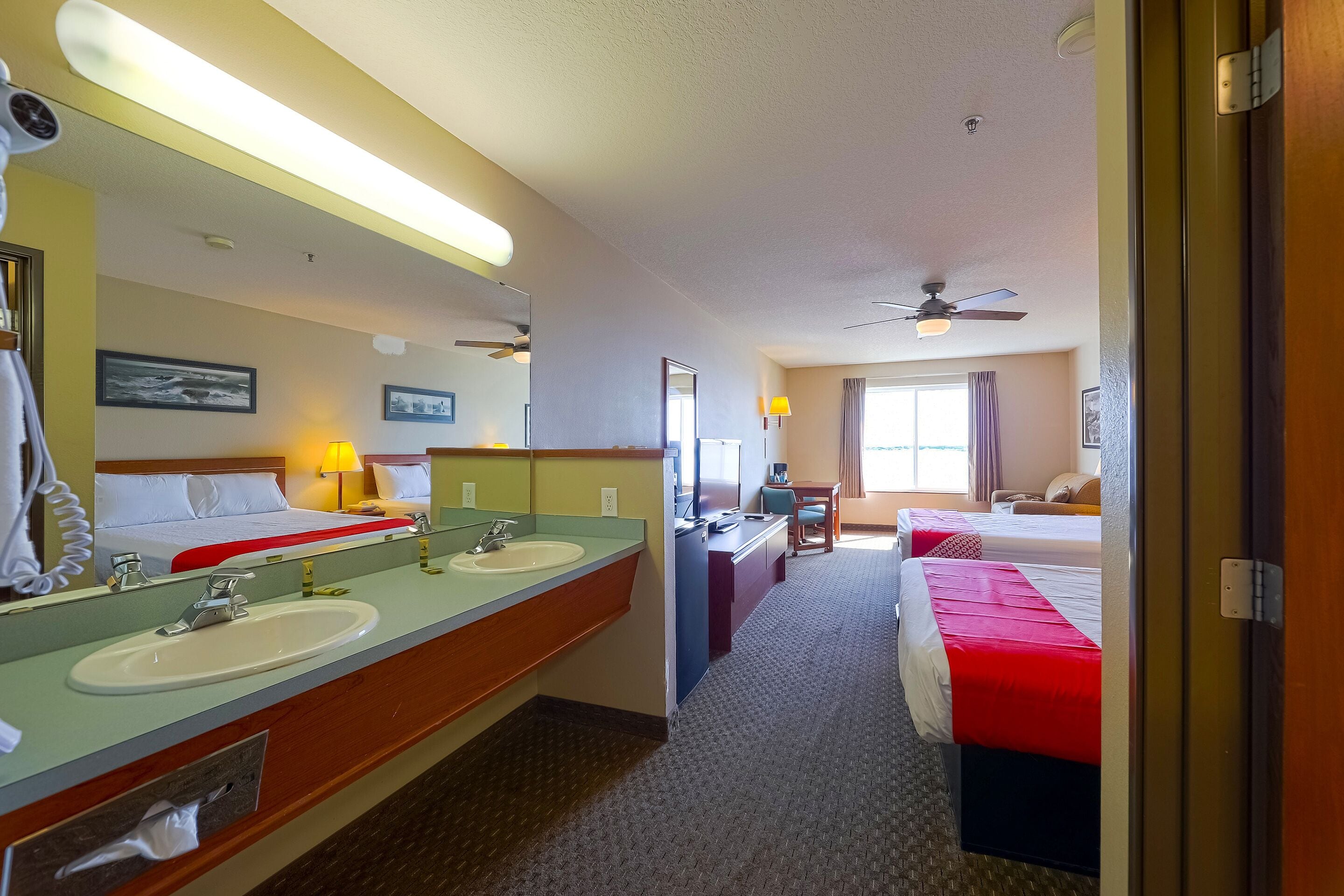 A hotel room with two beds, a bathroom with two sinks, a ceiling fan, and a large window.