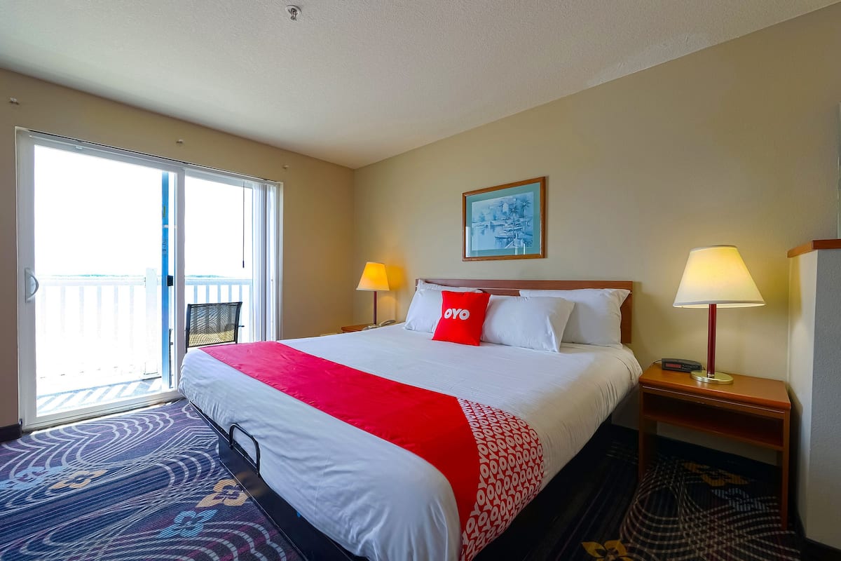 A hotel room with a bed, two bedside lamps, a red throw pillow, a framed picture on the wall, and a balcony with a view.