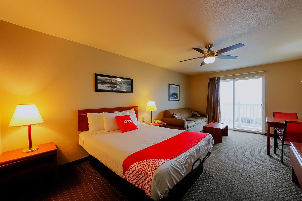 A hotel room with a bed, bedside tables, a desk, a chair, a ceiling fan, and a view of the outdoors.