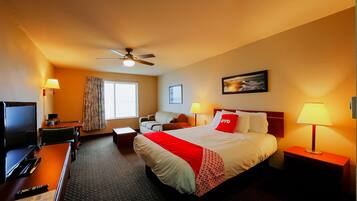 A hotel room with a bed, a desk with a computer, a TV, a ceiling fan, and a window with curtains.